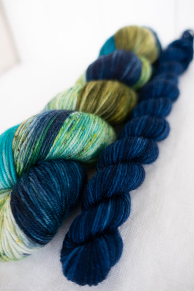 Skip Rope Yarn Co sock set - one 100g skein of speckled yarn in blue, green and olive with a 20 gram mini-skein in dark blue on a white background